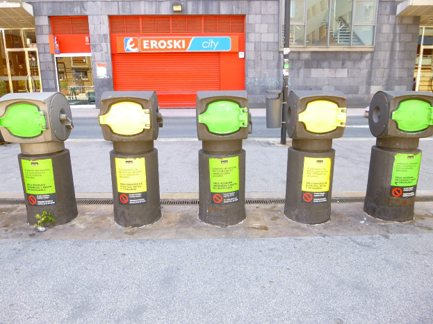 Reihe von Parkautomaten entlang einer Straße mit einem Gebäude mit Glasfenstern und einem Rollladen mit Text sowie einem nahen Müllcontainer.