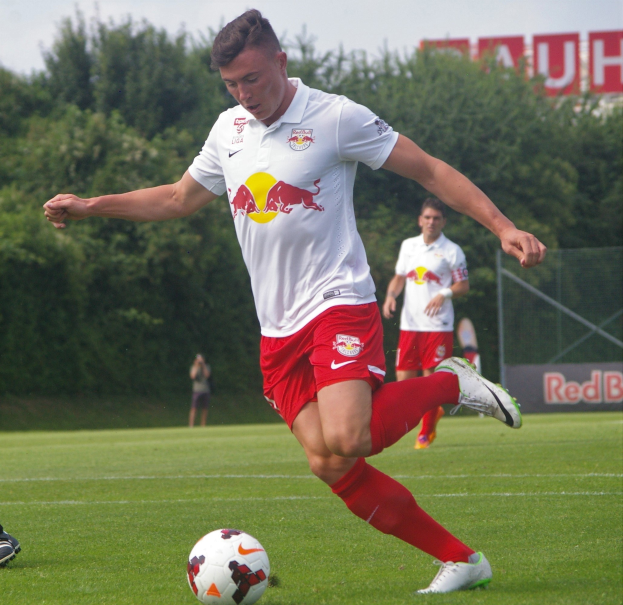 Ein professioneller Fußballspieler, der einen Fußball auf einem Rasenfeld kickt, mit "RB Leipzig" im Hintergrund und einem klaren blauen Himmel.