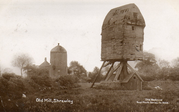 Alte hölzerne Mühle in schlechtem Zustand, umgeben von Bäumen und Pflanzen, mit sichtbarem Himmel im Hintergrund und Text unten.
