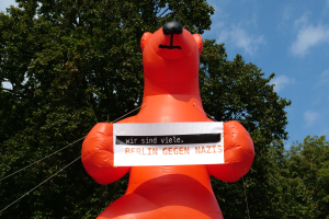 Aufblasbarer Bär mit einem Schild "Berlin Gegen Nazis" vor Bäumen und Wolken im Hintergrund.