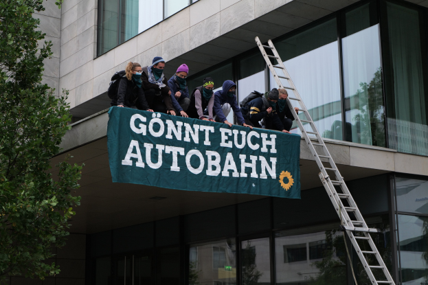 Gruppe von Menschen auf einem Gebäudedach mit einer Fahne, einem Baum links und Glasfenstern im Hintergrund, wahrscheinlich protestierend für die deutsche Regierung.