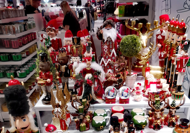 Ladeninneres voller Weihnachtsdekorationen, darunter Spielzeug, Tassen und andere Gegenstände auf Tischen und Regalen, mit Menschen, Lichtern und einer Wand im Hintergrund.