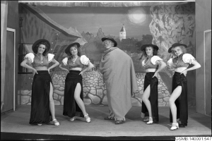 Fünf Frauen in Hüten stehen auf einer Bühne, lächeln und haben ein Gemälde und eine Tür im Hintergrund, schwarz-weiß, 1940er Jahre.