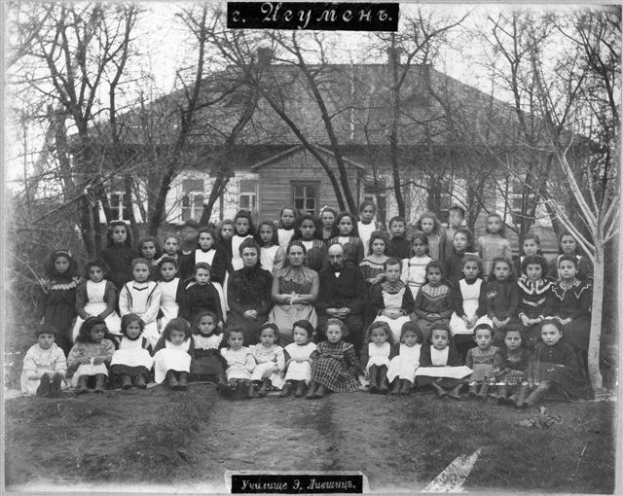 Schwarzes und weißes Foto von Kindern, die vor einem Gebäude mit Fenstern posieren, umgeben von Bäumen und einem klaren Himmel, mit Text unten, der 'Jugoslawien, die erste Schule der Welt' liest.