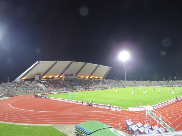 Ein großes Stadion voller Menschen, beleuchtet von Flutlicht, umgeben von Gras, Geländern, Stühlen und dem Himmel im Hintergrund.