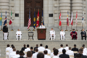 Eine Gruppe von Menschen in weißen Uniformen sitzt auf den Stufen vor einem Gebäude mit einem Podium, das von Fahnen flankiert ist.