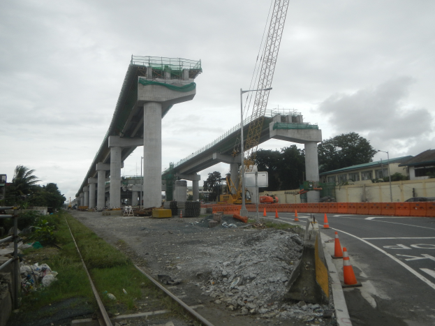 Eine Baustelle mit einer Brücke im Hintergrund, eine Straße mit Absperrbaken auf der rechten Seite, Steine und Gras auf dem Boden, eine Eisenbahnschiene auf der linken Seite, Bäume und Gebäude auf beiden Seiten der Straße und ein bewölkter Himmel.
