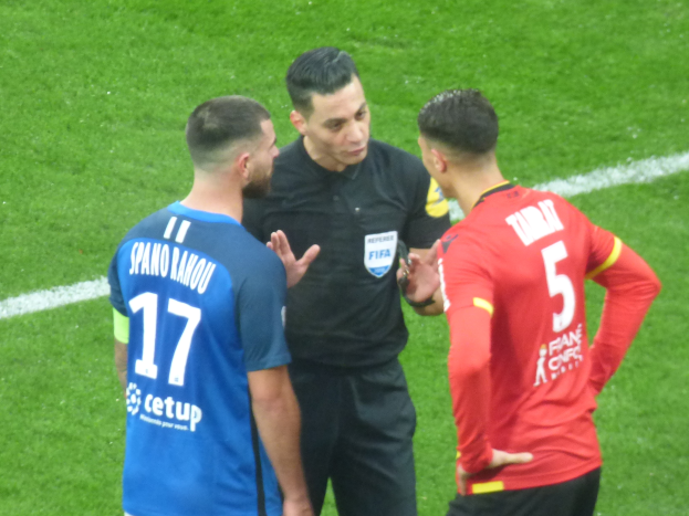 Ein Schiedsrichter in schwarzer Kleidung spricht mit zwei Männern auf einem Fußballfeld, einer in blauen und roten T-Shirts mit Aufschrift, beide tragen Brillen.