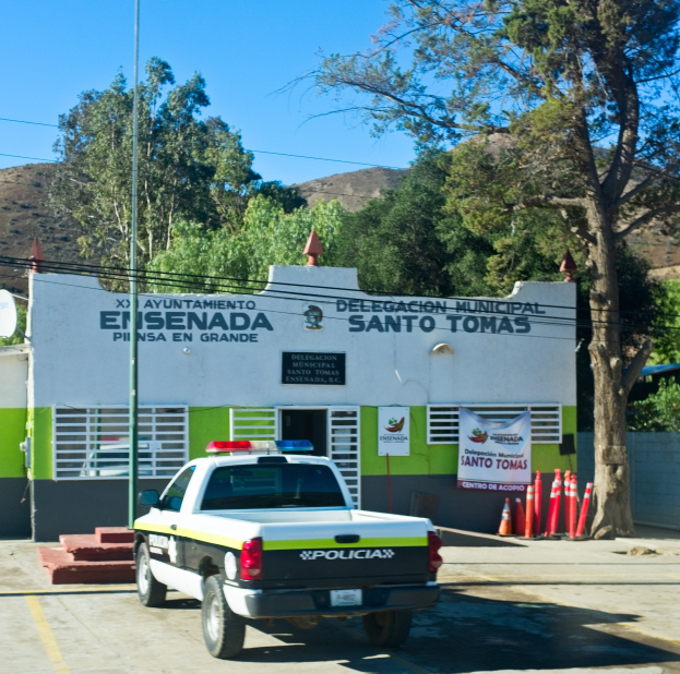 Polizeiauto vor einem Gebäude mit einem Schild, einer Fahne, Fenstern und einer Tür geparkt, mit Verkehrspfosten, einer Straßenlaterne, einer Satellitenschüssel, Drähten, Bäumen, Hügeln und einem bewölkten Himmel im Hintergrund.
