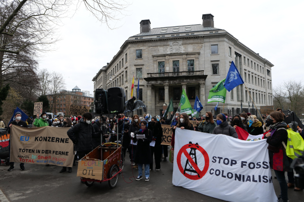 Eine große Gruppe von Menschen marschiert bei einer Protestaktion gegen fossile Brennstoffe, hält Schilder und Fahnen, mit einem Fahrzeug im Vordergrund und Gebäuden, Bäumen und einem klaren blauen Himmel im Hintergrund.