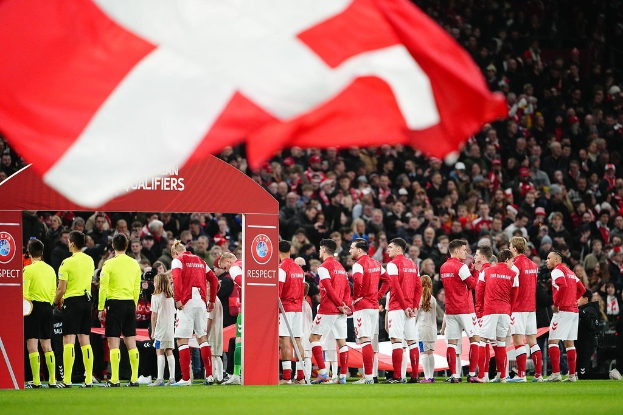 Eine Gruppe von Menschen auf einem Fußballfeld mit einer roten und weißen Flagge im Vordergrund, einem Bogen mit der Aufschrift "Bayern München vs. Bayern München Wett-Tipps & Vorschau" im Hintergrund und einer großen Menge im Stadion.