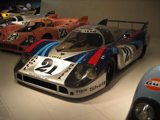 Ein Porsche 917K Le Mans Rennwagen in einem Museum ausgestellt, umgeben von anderen Sportwagen, mit Menschen, die zuschauen und einer Wand im Hintergrund, mit einem Textfeld auf der linken Seite.