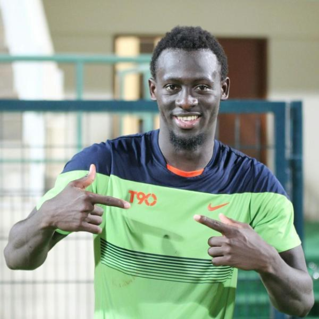 Ein Mann in einem grünen und blauen Hemd, der grinsend in die Kamera zeigt und im Hintergrund eine undeutliche Reling, Wand und Tür zu sehen sind.