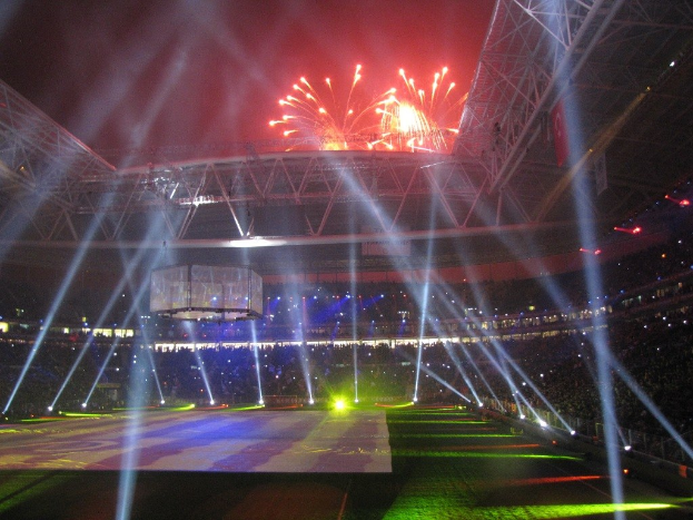 Olympisches Stadion in London bei Nacht, erleuchtet von hellen Lichtern und Feuerwerken, mit einer Menge drinnen und einem großen Bildschirm im Vordergrund.