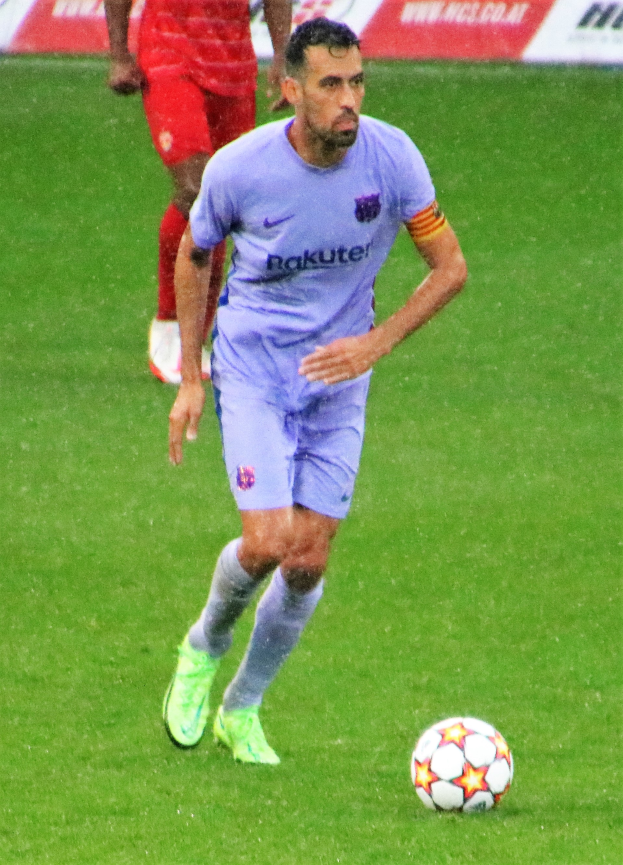 Ein Fußballspieler in Sportbekleidung und -schuhen ist im Einsatz auf dem Feld, wobei ein Textschild im Hintergrund sichtbar ist.