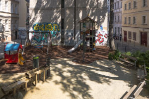 Spielplatz mit Graffiti, umgeben von Stadtgebäuden, Bäumen, Bänken und einem Mülleimer, mit Menschen und Fahrzeugen auf der Straße und Himmel im Hintergrund.