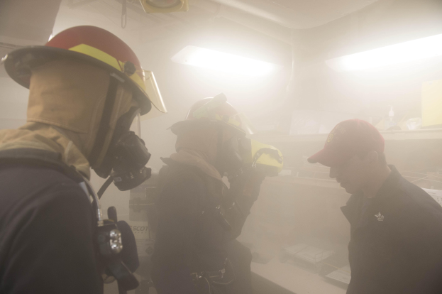 Feuerwehrleute in einem raucherfüllten Raum während einer Übung, einer hält eine Kamera, trägt Helme mit Rauch aus dem Mund.