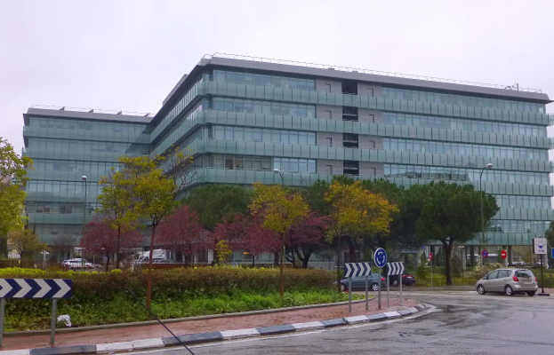 Großes Gebäude mit vielen Fenstern, der Hauptsitz der Europäischen Zentralbank, umgeben von Bäumen, Straßenlaternen, Fahrzeugen und Beschilderung unter einem bewölkten Himmel.