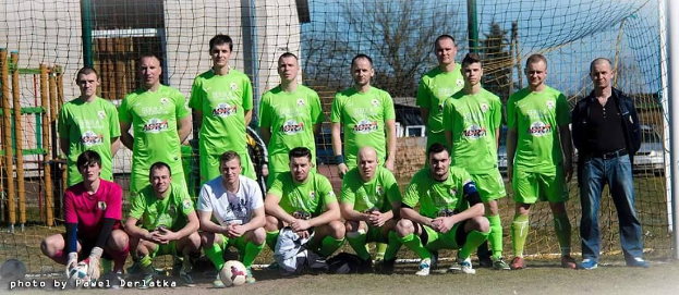 Eine Gruppe von Männern in grünen T-Shirts steht auf einem Fußballfeld, einige in Hockstellung, mit einem Ball davor und einem Torpfosten rechts daneben, umgeben von Bäumen, Gebäuden und Fahrzeugen unter einem Himmel.