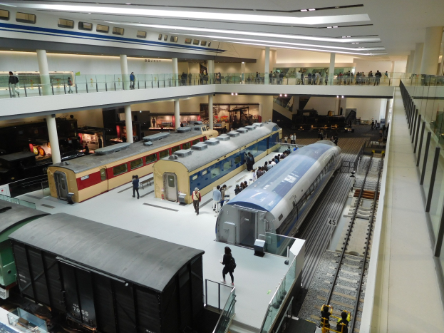 Eine große Museumsausstellung zeigt verschiedene Züge in unterschiedlichen Größen und Farben, mit Menschen, die vom Boden, Geländern und Säulen unter Deckenbeleuchtung beobachten.