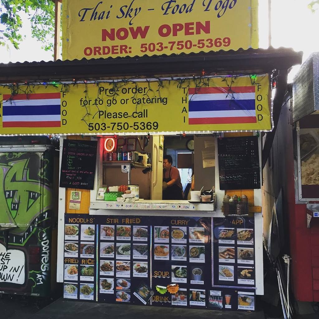 Läden, Getränkedosen, Speisekarten, eine Uhr, Gewürze in Plastikbehältern, Würzmittel, Werbetafeln, Namensschilder und Himmel.