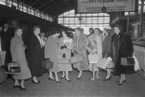 Schwarzes und weißes Bild von Frauen, die an einem Bahnhof stehen, links ein Zug und rechts Geländer und eine Tafel, jede hält eine Tasche.