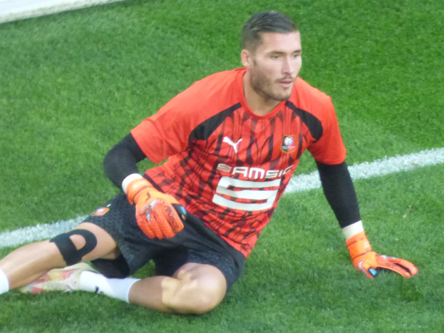 Fußballspieler in rotem T-Shirt, schwarzen Shorts, weißen Socken und orangen Baseball-Handschuhen auf dem Boden sitzend.