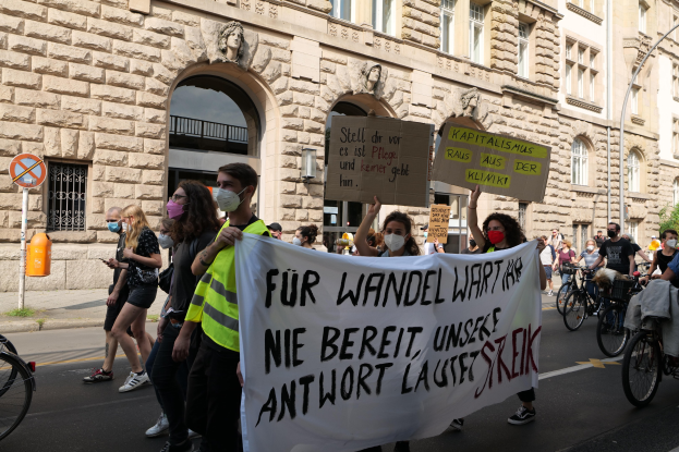 Eine Gruppe von Menschen bei einer Protestdemo auf der Straße, einige halten Schilder und andere fahren Fahrräder, im Hintergrund ein Gebäude mit Fenstern, Bögen, Säulen und Skulpturen.