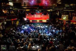 Eine große Menschenmenge sitzt auf Stühlen vor einer Bühne mit Bildschirmen und Bannern, beleuchtet von Scheinwerfern und umgeben von Geländern, mit einem Wasserzeichen in der linken unteren Ecke, und wartet auf ein Online-Marketing-Rockstars-Event.