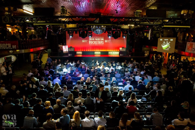 Eine große Menschenmenge sitzt auf Stühlen vor einer Bühne mit Bildschirmen und Bannern, beleuchtet von Scheinwerfern und umgeben von Geländern, mit einem Wasserzeichen in der linken unteren Ecke, und wartet auf ein Online-Marketing-Rockstars-Event.