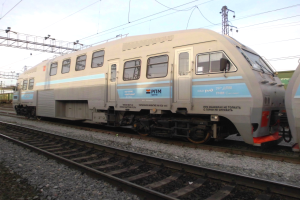 Ein Diesellokomotive fährt auf Schienen neben einem anderen Zug, mit Strommasten und Kabeln im Hintergrund unter einem klaren blauen Himmel.