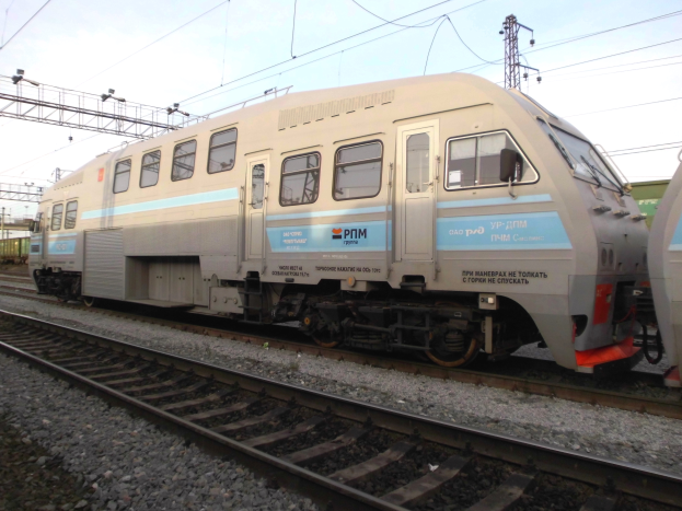 Ein Diesellokomotive fährt auf Schienen neben einem anderen Zug, mit Strommasten und Kabeln im Hintergrund unter einem klaren blauen Himmel.