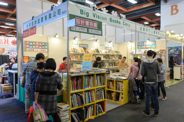 Eine Gruppe von Menschen, die Bücher auf einem Messestand mit Regalen voller Bücher, Informationsschildern und Deckenbeleuchtung durchstöbert.