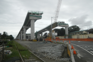 Baustelle mit einer Brücke im Hintergrund, Straße durch Verkehrskegel markiert, Steine und Gras am Boden, Eisenbahnschiene links, Bäume und Gebäude auf beiden Seiten und bewölkter Himmel oben.