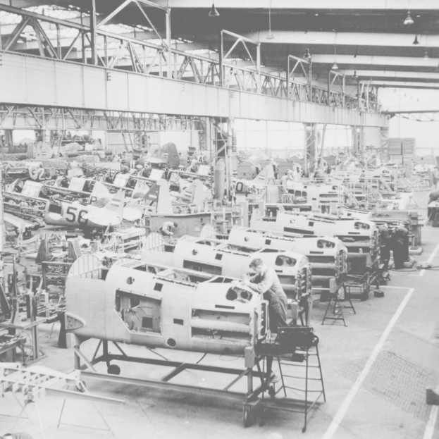 Ein Schwarz-Weiß-Foto einer Fabrik, die voller Flugzeuge ist, mit Arbeitern, die an Maschinen auf dem Boden arbeiten, und Lampen an der Decke.