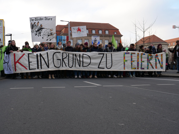 Demonstranten mit einem Banner "Kein Grund zu Feiern" gegen deutsche Sparmaßnahmen, mit Gebäuden, Bäumen und einem klaren Himmel im Hintergrund.