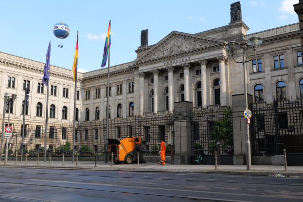 Großes modernes Gebäude mit zahlreichen Fenstern, identifiziert als der Bundestag in Berlin, Deutschland, umgeben von Straßeninfrastruktur, Füßgängern und Flaggen, mit einem bewölktem Himmel im Hintergrund.