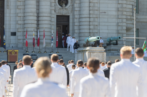 Eine Gruppe von Menschen in weißen Uniformen und Mützen steht vor einem Gebäude mit Säulen und einer Tür, mit Fahnen, einem Podium mit Mikrofon, Stufen und Kanonen im Hintergrund, was auf eine Abschlussfeier der Marineakademie hindeutet.