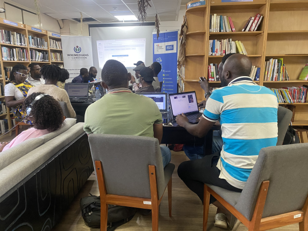 Eine Gruppe von Menschen, die um einen Tisch mit Laptops in einer Bibliothek sitzen, umgeben von Bücherregalen, mit einem Banner, Bildschirm, Deckenleuchten und Taschen auf dem Boden.