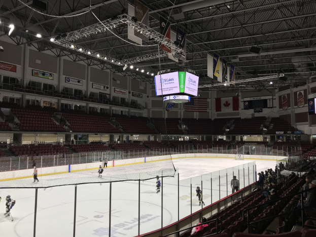 Hockey-Spiel in einer großen Arena mit einem zentralen Netz, Zuschauern auf Stühlen, Deckenbeleuchtung und Trägern sowie Bannern an den Wänden, die "St. Louis Blues Arena" beschriftet sind.