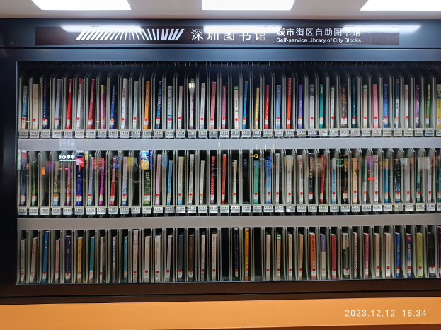 Eine gut beleuchtete Bibliothek mit hohen Bücherregalen voller Bücher, die ein Schild mit Text und ein Wasserzeichen am Boden zeigt, ähnlich einer Selbstbedienungsbibliothek der Stadtviertel.