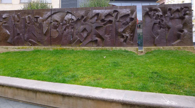 Große Metallskulptur der Berliner Mauer in einer grünen Fläche umgeben von Bäumen und Gebäuden mit Fenstern im Hintergrund.