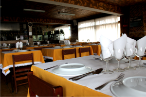 Ein Restaurantinnenraum mit für ein Essen gedeckten Tischen, mit Tellern, Besteck, Gläsern und Servietten, einem Fenster mit Vorhängen im Hintergrund, Deckenleuchten und Text am unteren Bildrand.