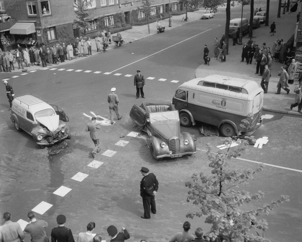 Schwarz-weißes Bild eines Autounfalls an einer Straßenkreuzung mit einem Lieferwagen, anderen Fahrzeugen und Menschen, die sich darum versammeln, mit Gebäuden, Bäumen, Pfählen und Texttafeln im Hintergrund.