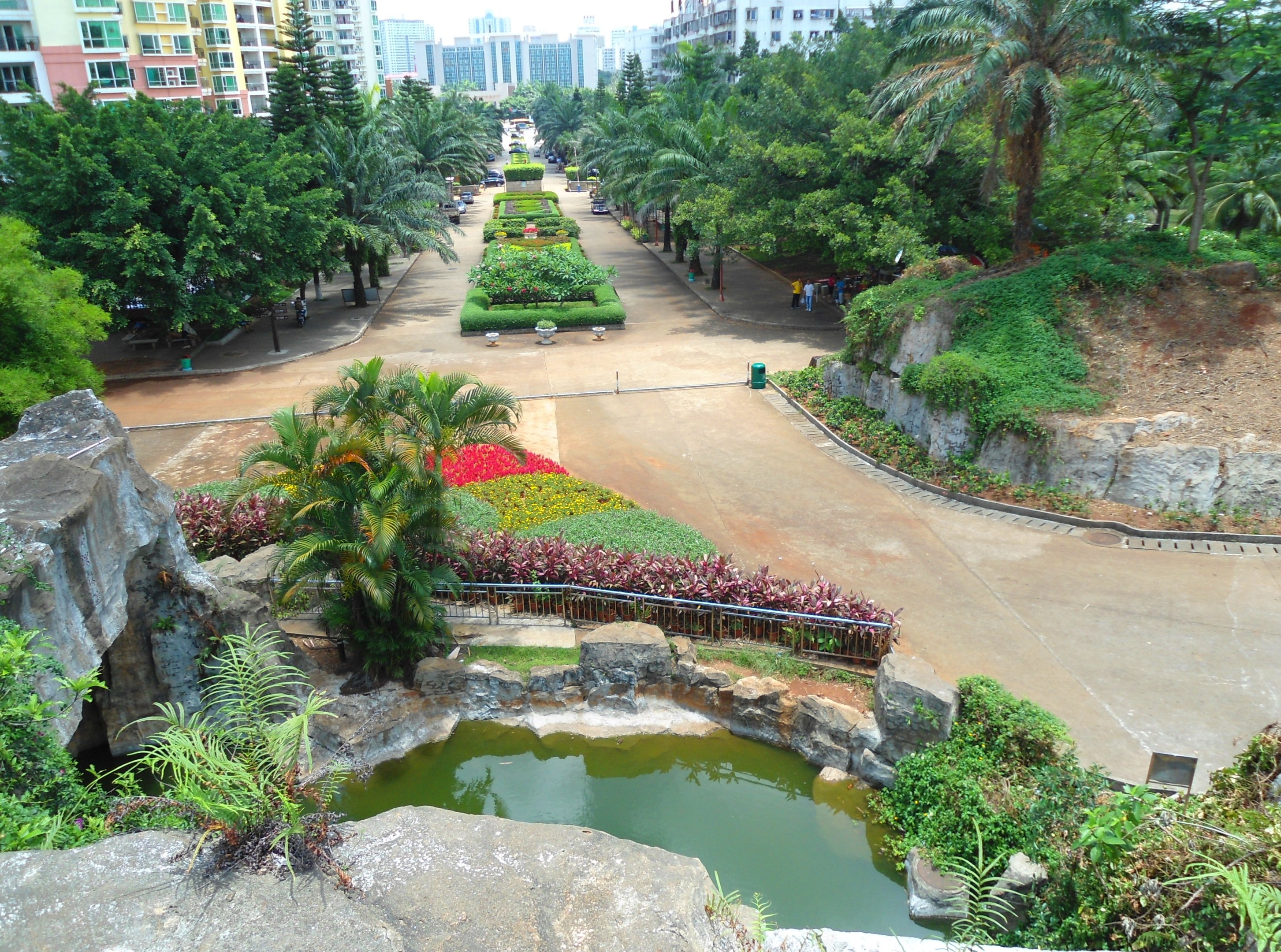 Luftbild eines Parks mit einem Teich, umgeben von Felsen, Grünflächen und Blumen, mit Gebäuden und einem klaren blauen Himmel im Hintergrund, mit Menschen, die herumlaufen.