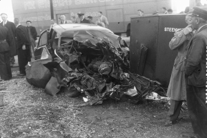 Eine Schwarz-Weiß-Aufnahme eines auf der Seite liegenden Wagens mit mehreren Menschen drumherum, einige tragen Mützen, und einem Lkw im Hintergrund.