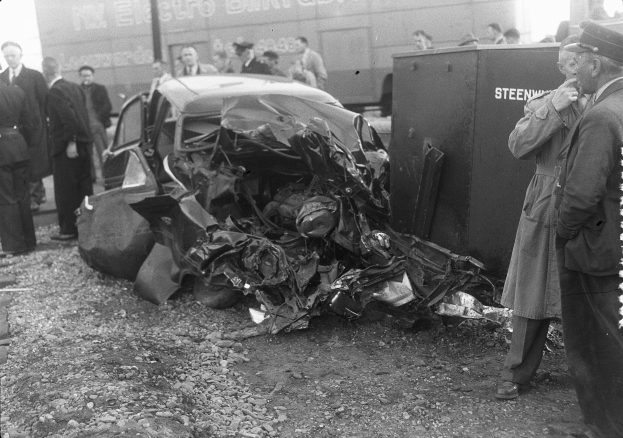 Eine Schwarz-Weiß-Aufnahme eines auf der Seite liegenden Wagens mit mehreren Menschen drumherum, einige tragen Mützen, und einem Lkw im Hintergrund.