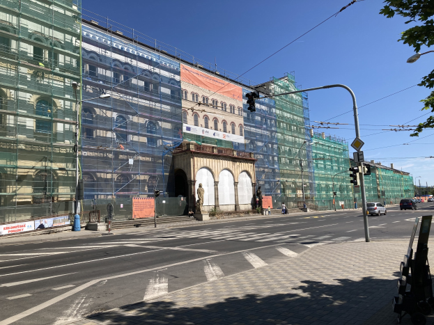 Großer mehrstöckiger Bau unter construction in Berlin mit sichtbarem Gerüst, umgeben von städtischer Infrastruktur wie Straßenlaternen, Ampeln, Fahrzeugen, Fußgängern, Bäumen und Werbetafeln unter einem bewölkten Himmel.