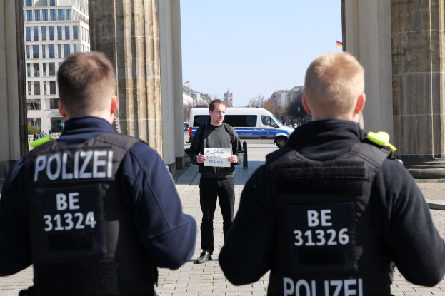 Drei Polizeibeamte stehen vor einem Gebäude, mit einem Mann, der ein Papier zwischen ihnen hält, flankiert von Säulen und einem belebten städtischen Hintergrund.