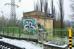 Eine Bahnstrecke mit Graffiti an der Seite, umgeben von Gelöndern, Pfosten, Lichtern und Bäumen, mit Törmen und Dröhnen im Hintergrund und Schnee am Boden.